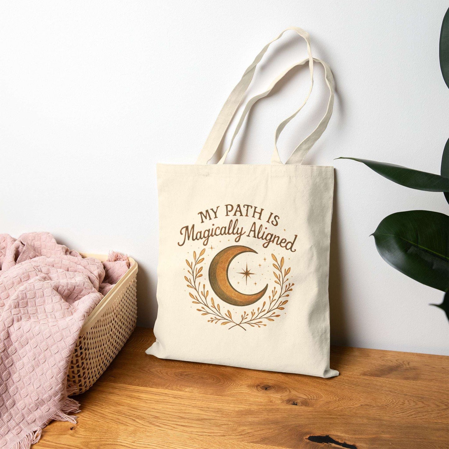Boho Moon Canvas Tote with 'My Path Is Magically Aligned' quote, resting on wooden table beside pink towel and plant. Ideal for spiritual or bohemian lifestyles.