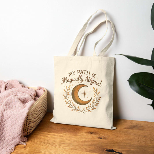 Boho Moon Canvas Tote with 'My Path Is Magically Aligned' quote, resting on wooden table beside pink towel and plant. Ideal for spiritual or bohemian lifestyles.