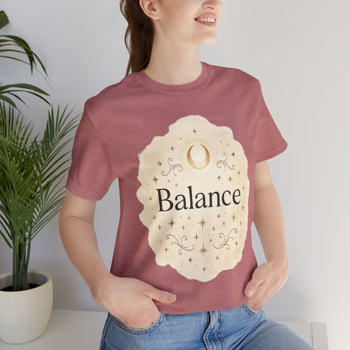 Person wearing a maroon t-shirt with 'Balance' design, standing indoors next to a plant.