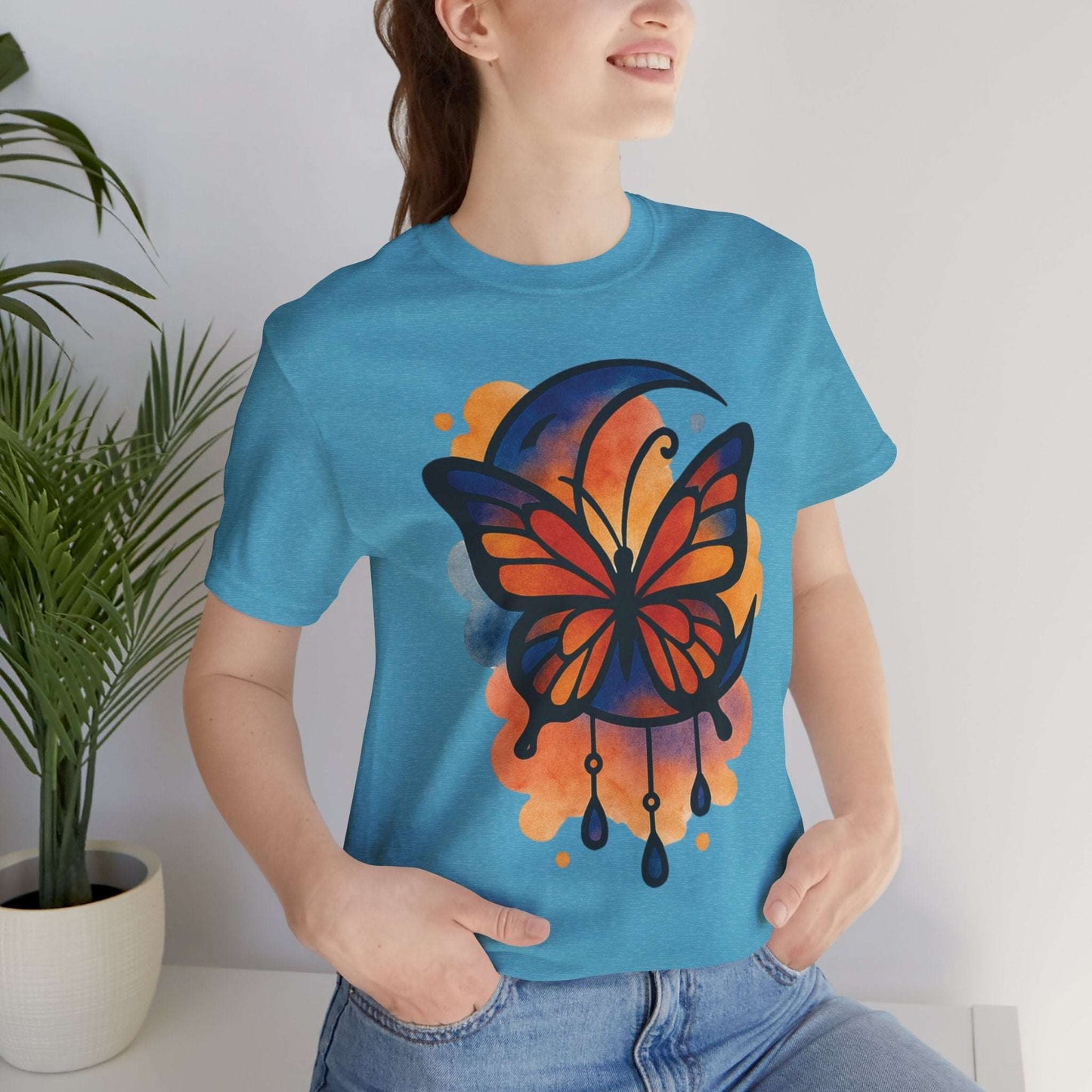 Person wearing a blue t-shirt with a colorful butterfly design, standing indoors next to a plant.