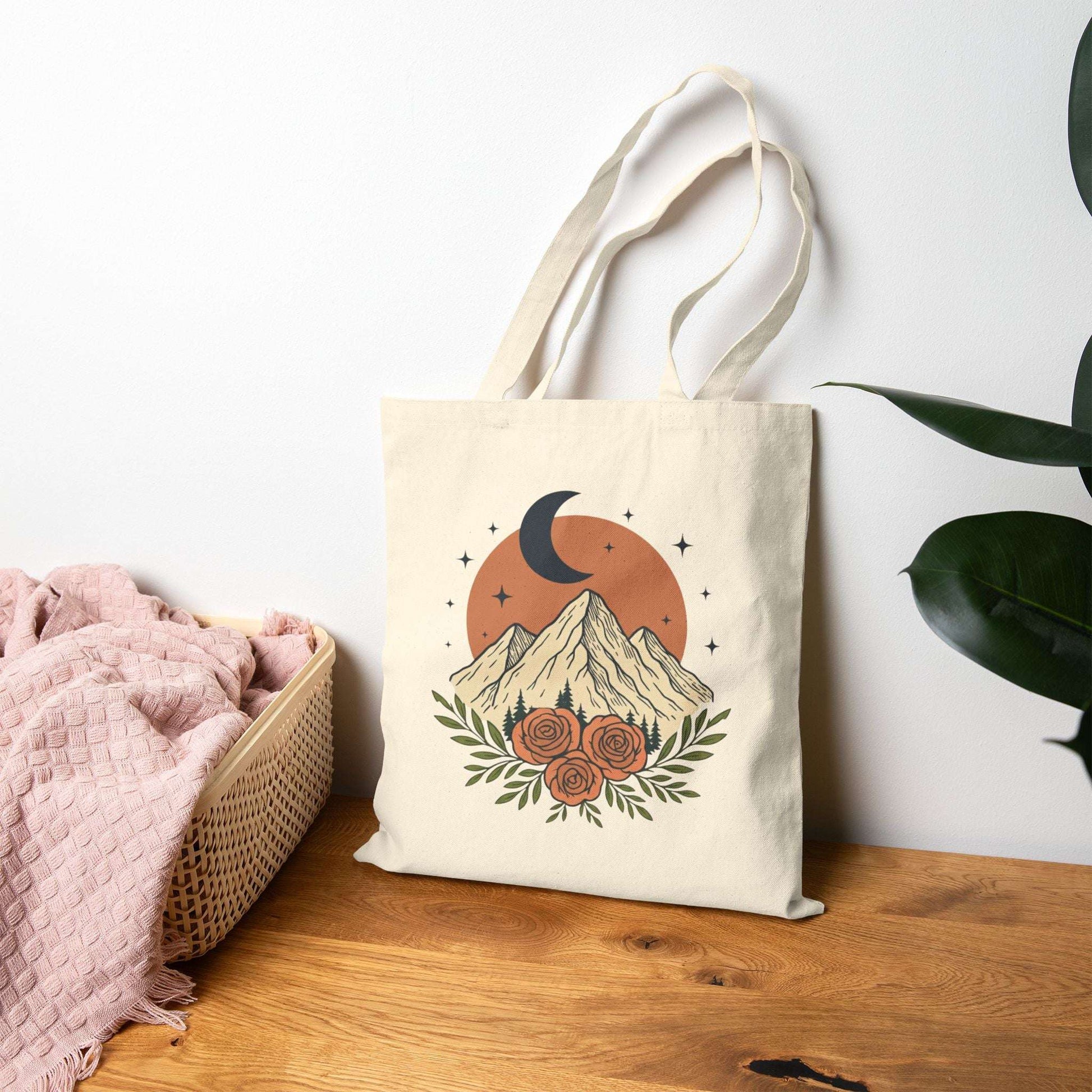 Canvas tote bag with vintage mountain, moon, and rose design resting against a wall on a wooden surface. Eco-friendly shopper for daily use.
