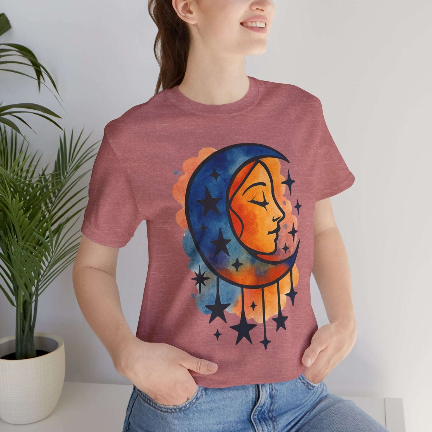 Person wearing a t-shirt with a colorful moon and star design, standing indoors next to a plant.