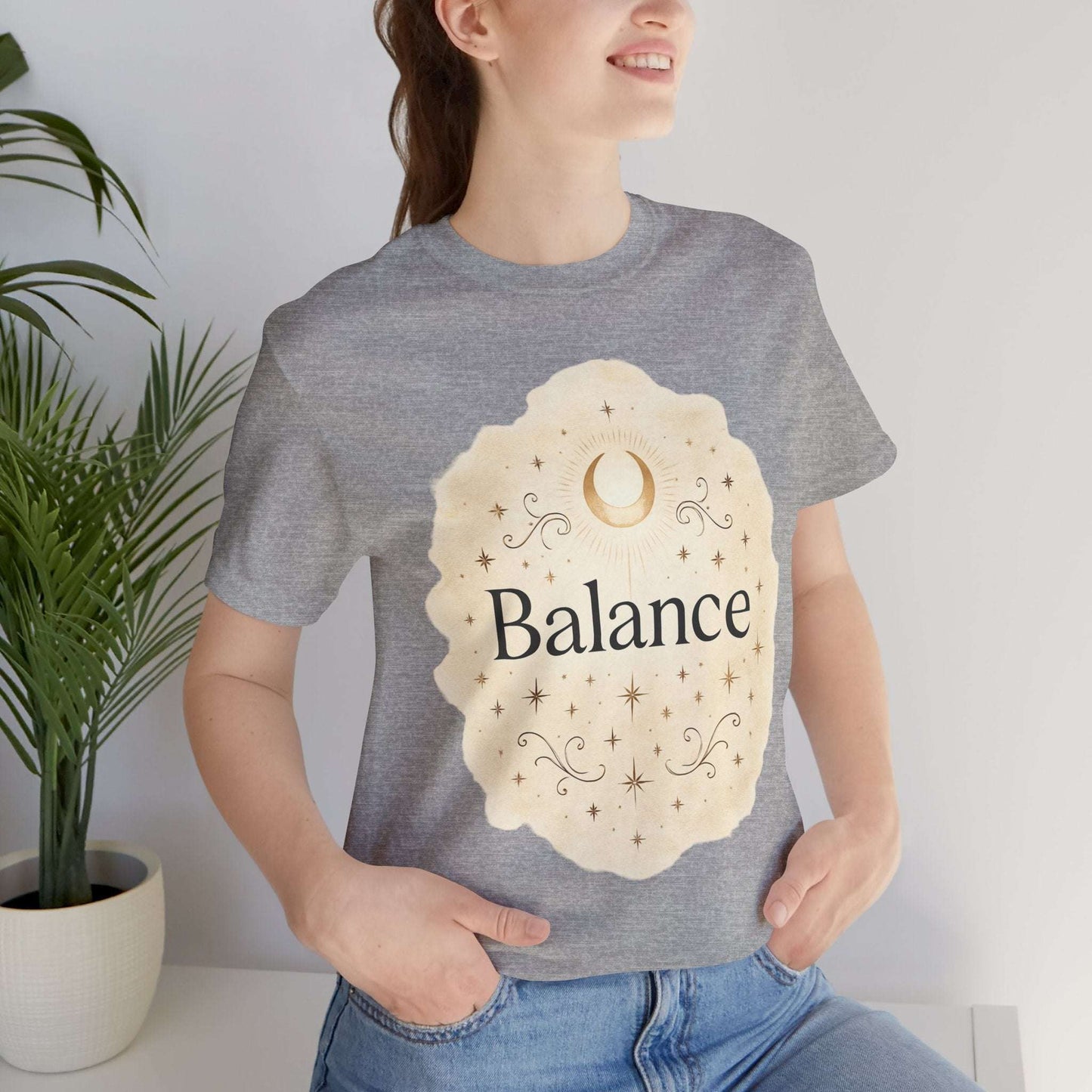 Person wearing a gray t-shirt with 'Balance' design, standing indoors with a plant in the background.