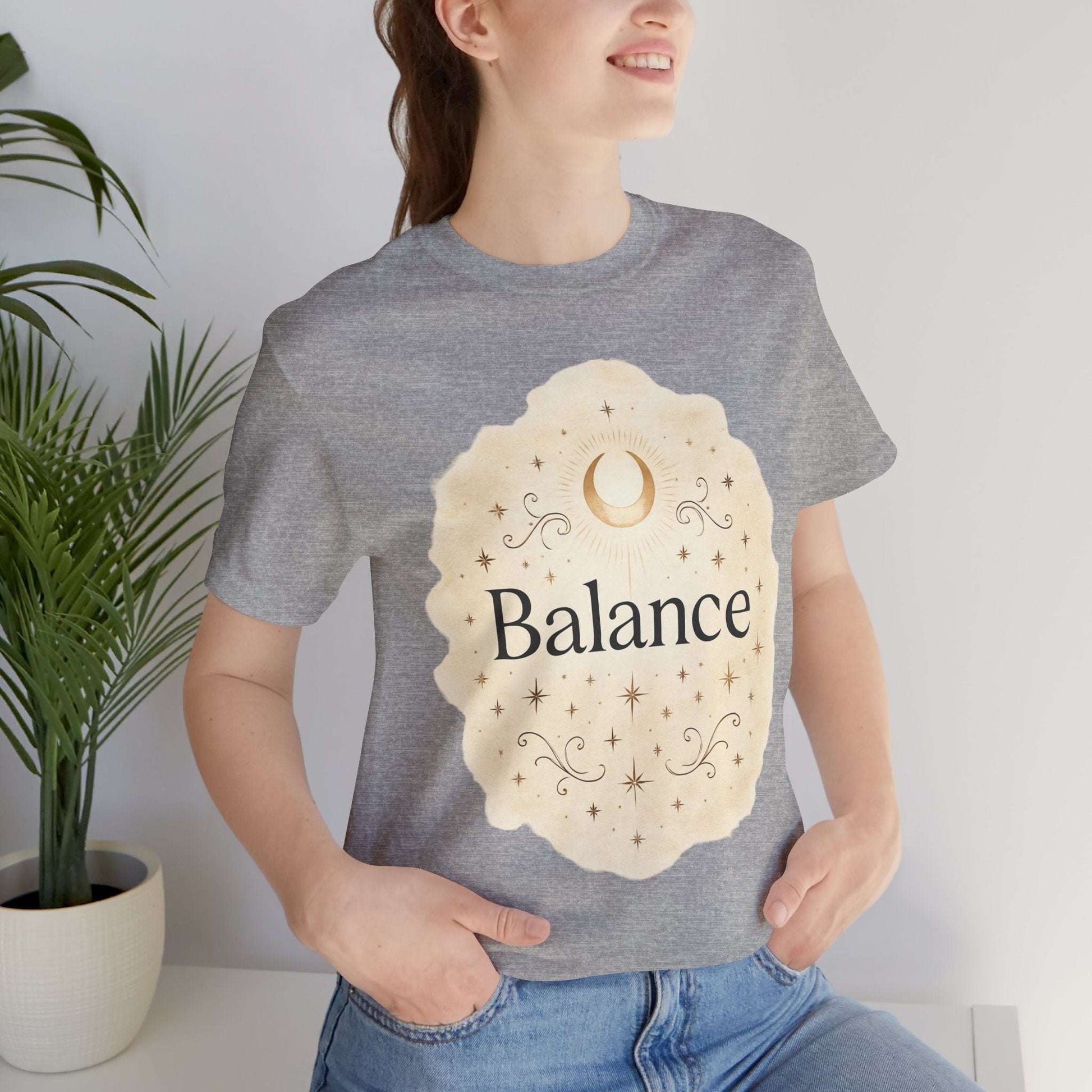 Person wearing a gray t-shirt with 'Balance' design, standing indoors with a plant in the background.