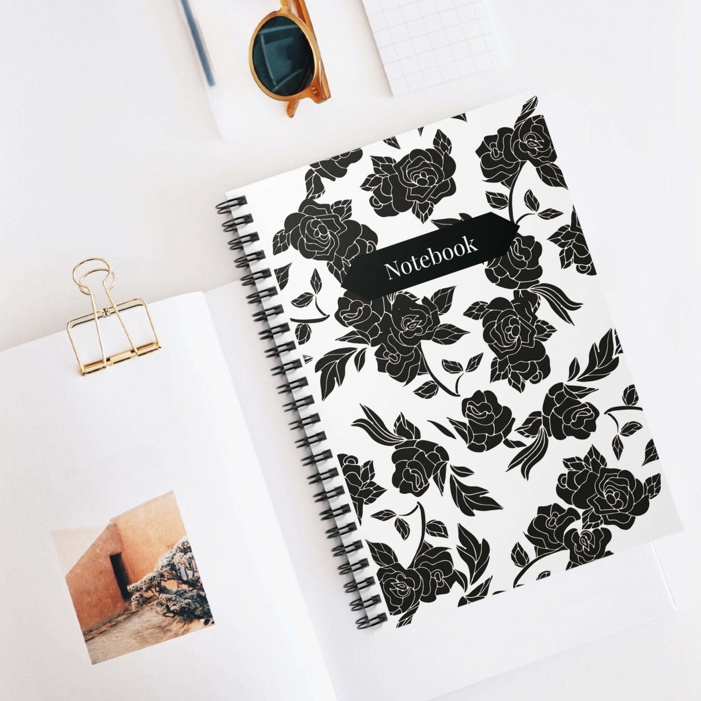 Notebook with black floral design on a white surface with sunglasses and a photo clip.