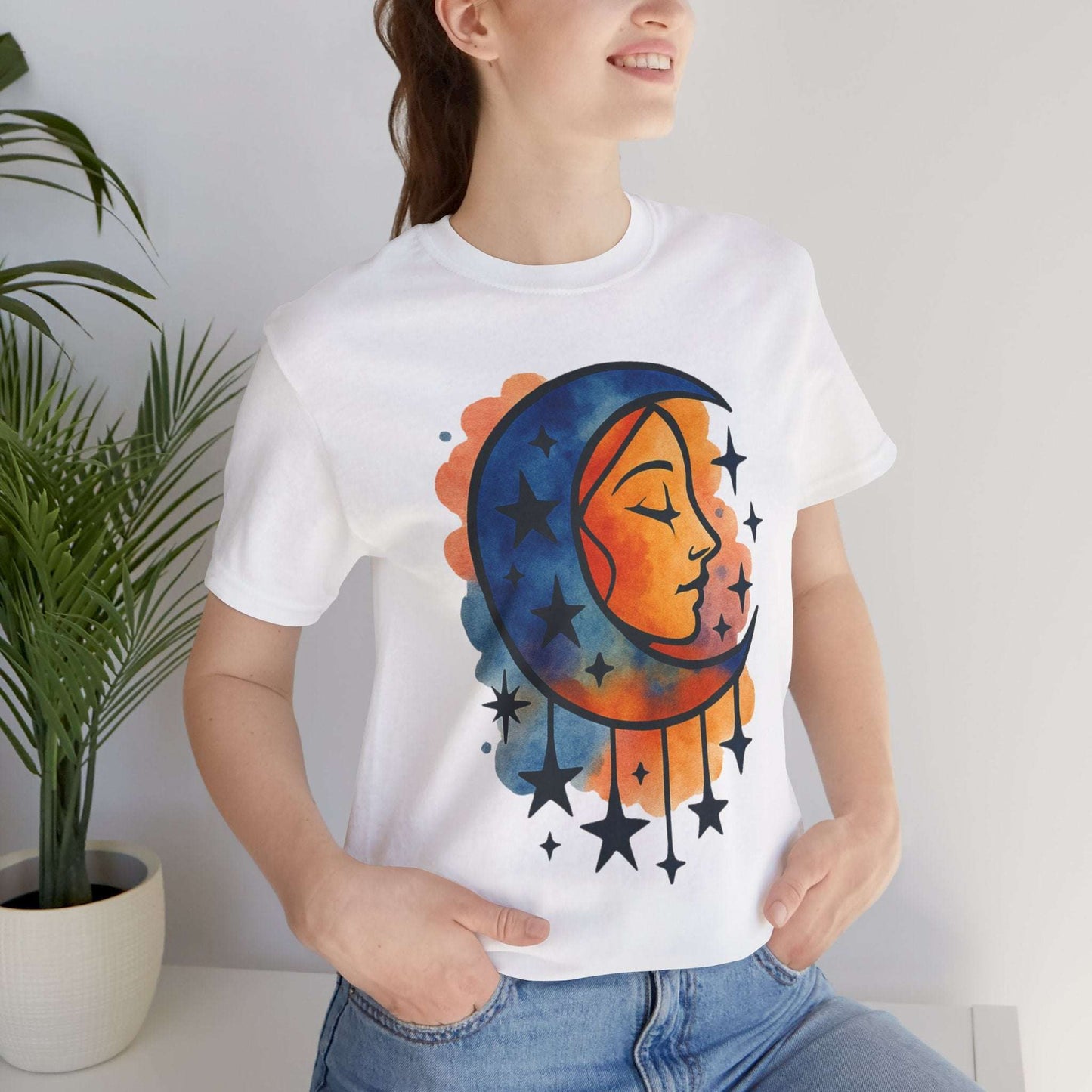 Person wearing a white t-shirt with a colorful moon and star design, standing indoors next to a plant.