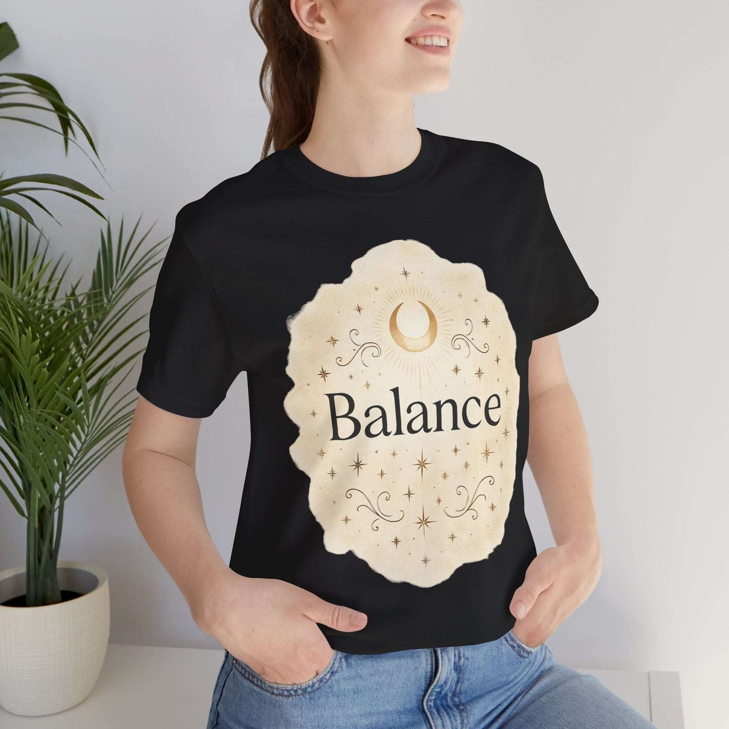 Person wearing a black t-shirt with 'Balance' design, standing indoors with a plant in the background.