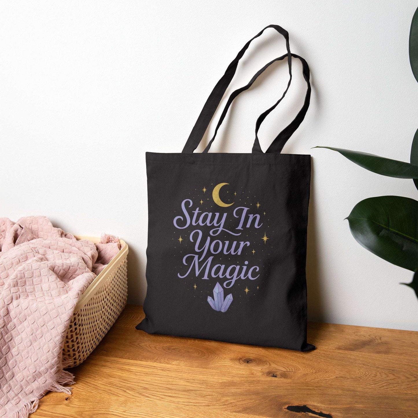 Black canvas tote bag featuring the quote 'Stay In Your Magic', displayed on a chair beside a plant, lifestyle product photo.